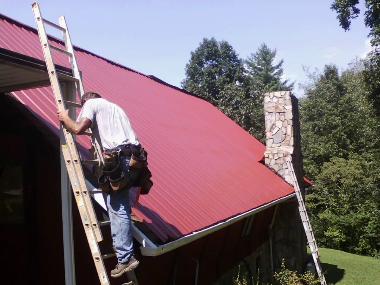 Roofer on a ladder beside a red metal roof and stone chimney in Trenton